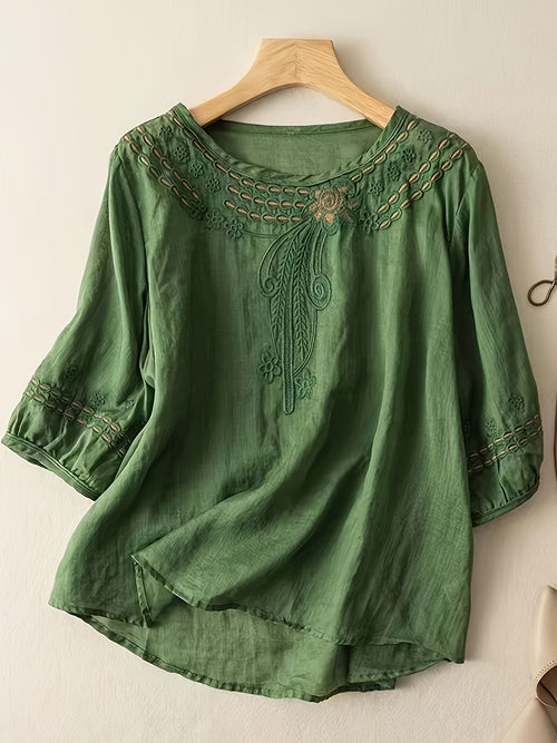 Green embroidered blouse on a wooden hanger against a beige wall.